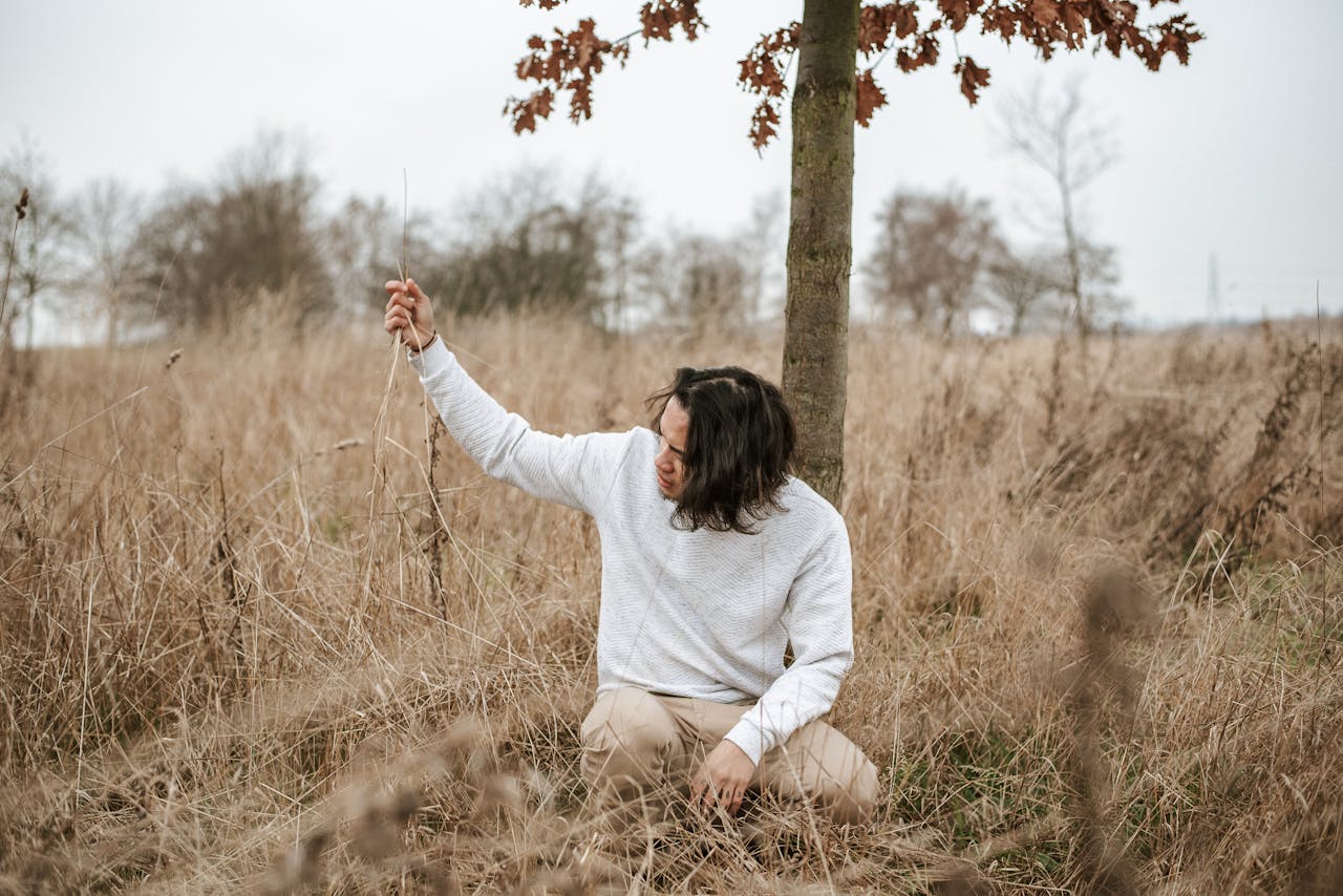 Depressed lonely male sitting among dry grass in autumn meadow in cloudy day