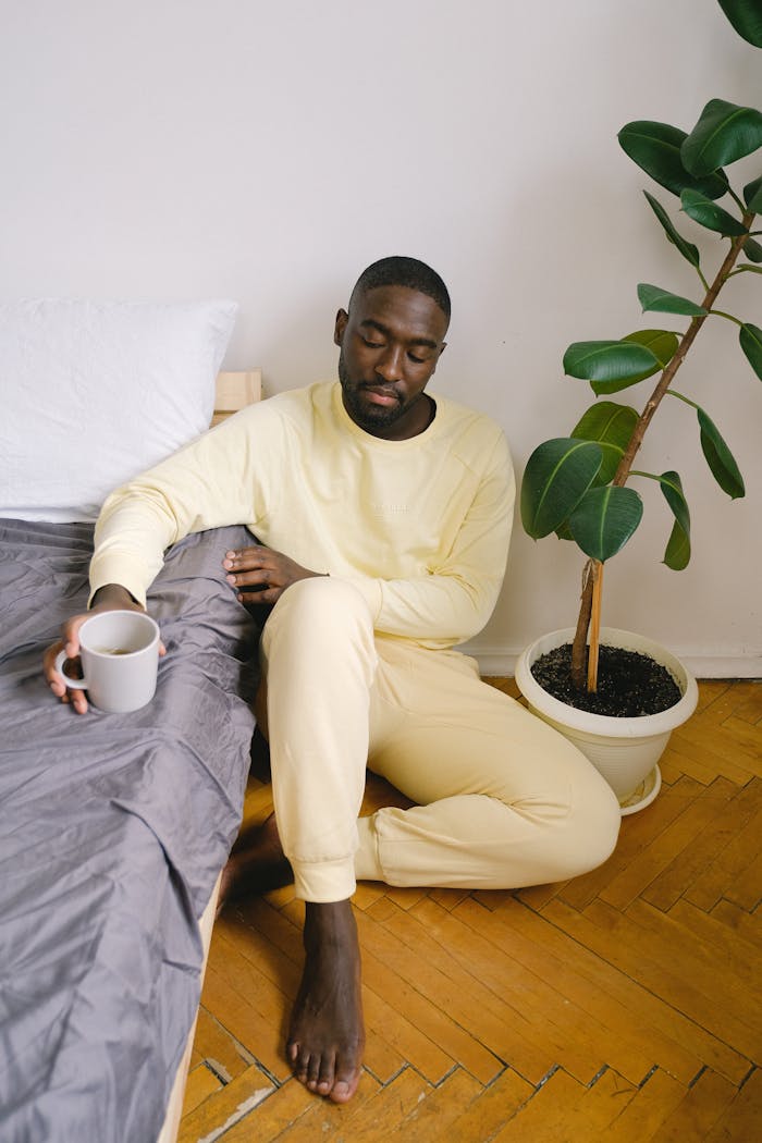 Sad African American male in pajamas sitting on floor with crossed legs and mug of hot drink at home